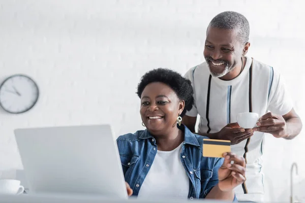 Mujer afroamericana mayor sosteniendo tarjeta de crédito cerca de la computadora portátil y marido feliz con taza - foto de stock