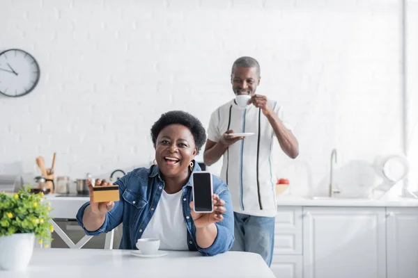 Feliz adulto mayor afroamericana mujer sosteniendo tarjeta de crédito y teléfono inteligente con pantalla en blanco cerca del marido - foto de stock