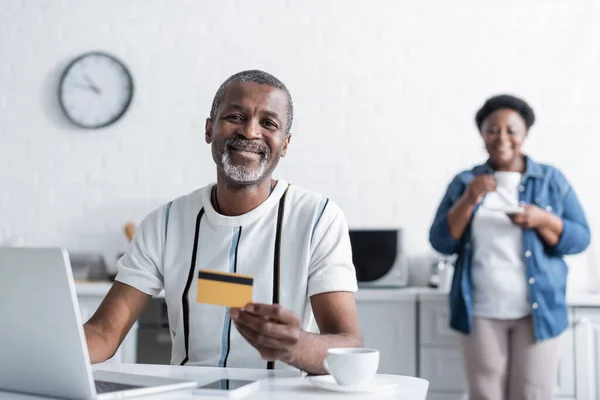 Feliz homem americano africano sênior segurando cartão de crédito perto de laptop e esposa borrada na cozinha — Fotografia de Stock