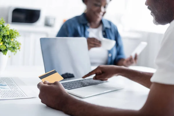 Vista cortada do homem americano africano sênior segurando cartão de crédito e usando laptop perto da esposa turva — Fotografia de Stock