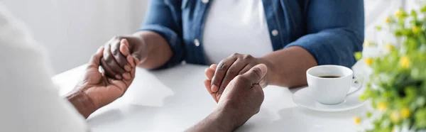 Vista recortada de la pareja afroamericana de alto nivel tomados de la mano cerca de la taza de café, pancarta - foto de stock