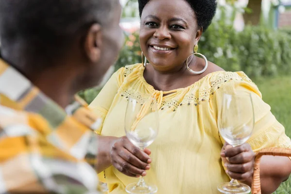 Happy african american woman smiling and holding wine glasses near husband during picnic — Stock Photo