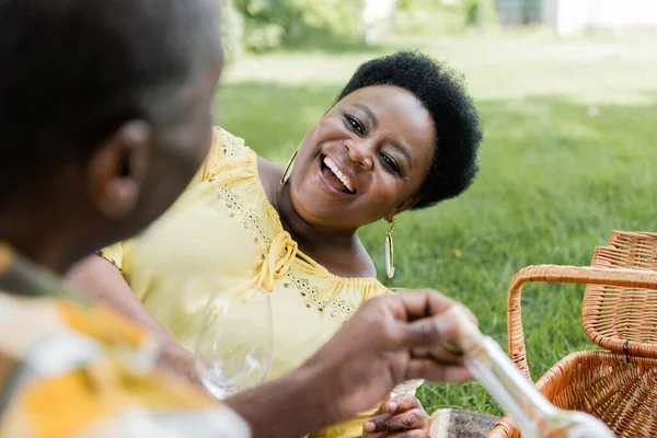 Возбужденная африканская американка улыбается и держит бокал вина рядом с мужем во время пикника — стоковое фото