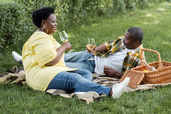 Alegre y senior africano americano pareja celebración copas de vino durante picnic - foto de stock