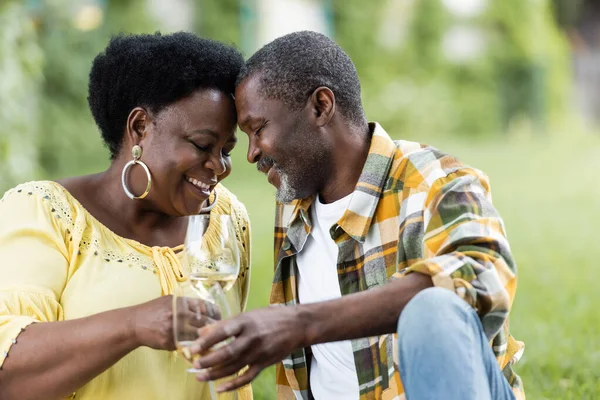 Tierna y mayor pareja afroamericana sonriendo mientras sostiene vasos de vino - foto de stock