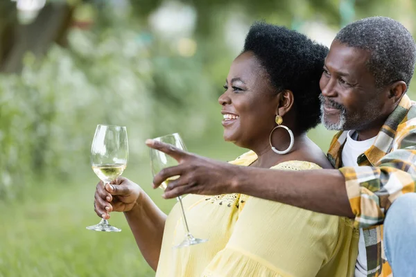 Romantic senior african american man pointing away near happy wife with glass of wine — Stock Photo