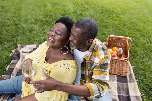 Vista de alto ángulo de alegre y senior africano americano pareja tintineo vasos de vino durante el picnic en el parque - foto de stock