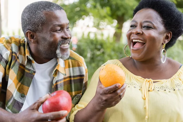 Cheerful and senior african american couple holding fruits during picnic — Stock Photo