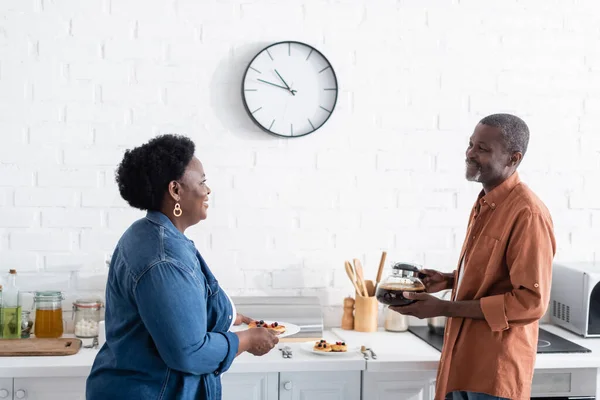 Glückliches älteres afrikanisch-amerikanisches Paar schaut einander an, während es Pfannkuchen und Kaffeekanne in der Küche hält — Stockfoto