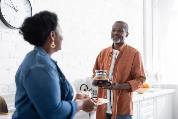 Alegre casal africano sênior americano olhando um para o outro enquanto segurando panquecas e cafeteira na cozinha — Fotografia de Stock