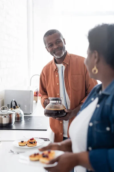 Gai senior afro-américain tenant cafetière et regardant femme — Photo de stock