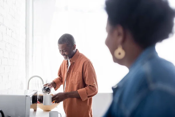 Alegre y sénior afroamericano hombre sosteniendo cafetera cerca de taza y esposa en primer plano borrosa - foto de stock