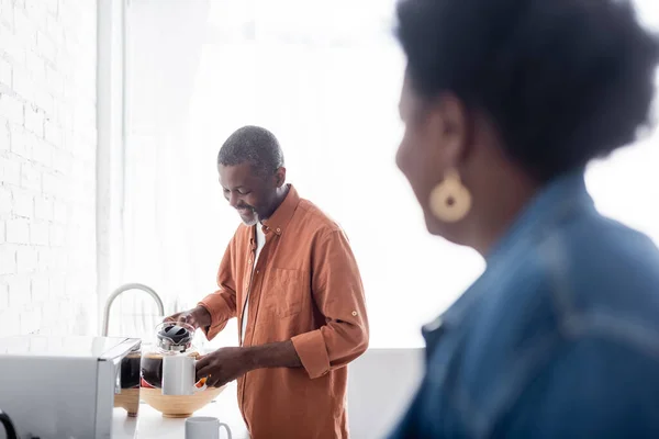 Cheerful and senior african american man holding coffee pot near cup and wife on blurred foreground — Stock Photo