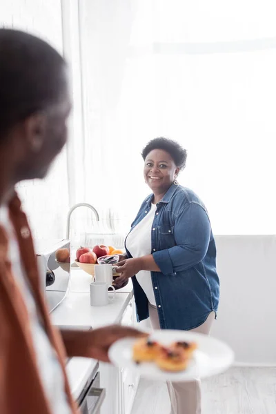 Fröhliche und ältere afrikanisch-amerikanische Frau mit Kaffeekanne in der Nähe von Tassen und verschwommene Frau — Stockfoto