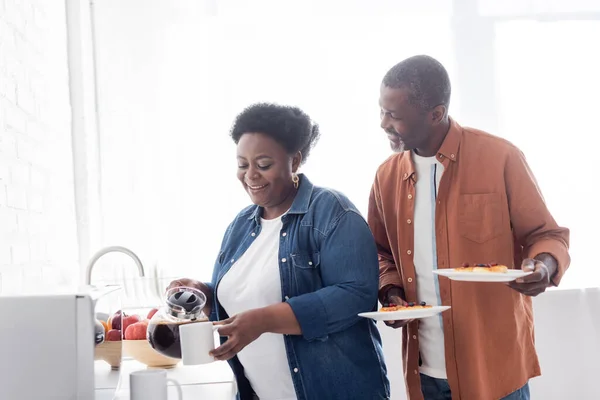 Femme afro-américaine gaie et aînée versant du café dans une tasse près du mari — Photo de stock