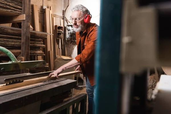 Mature carpenter cutting board on circular saw on blurred foreground — Stock Photo