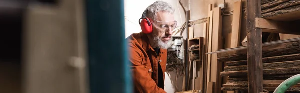 Bearded woodworker in earmuffs and goggles in workshop on blurred foreground, banner — Stock Photo