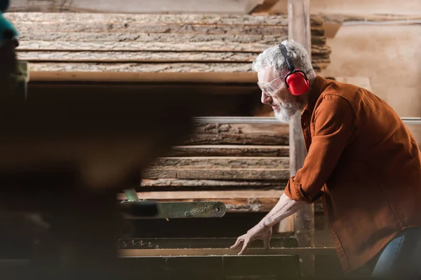 Side view of carpenter in protective earmuffs working on circular saw on blurred foreground — Stock Photo