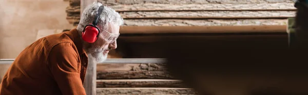 Vue latérale du menuisier dans des cache-oreilles de protection près des planches en atelier de menuiserie, bannière — Photo de stock