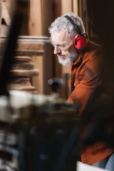 Menuisier barbu dans des lunettes et des cache-oreilles protecteurs travaillant sur le premier plan flou — Photo de stock