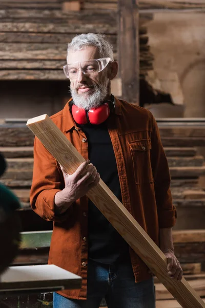Mature furniture designer in goggles holding wooden plank in carpentry studio — Stock Photo
