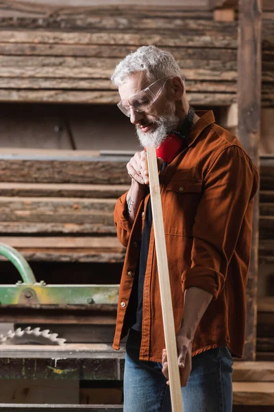 Diseñador de muebles de pelo gris en gafas comprobar tablón de madera en taller de carpintería - foto de stock