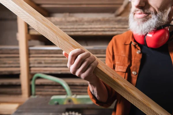 Vue recadrée d'un charpentier barbu tenant une planche de bois dans un atelier de menuiserie — Photo de stock