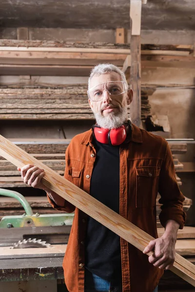 Бородатый деревообработчик с доской, смотрящий в камеру в мастерской — стоковое фото