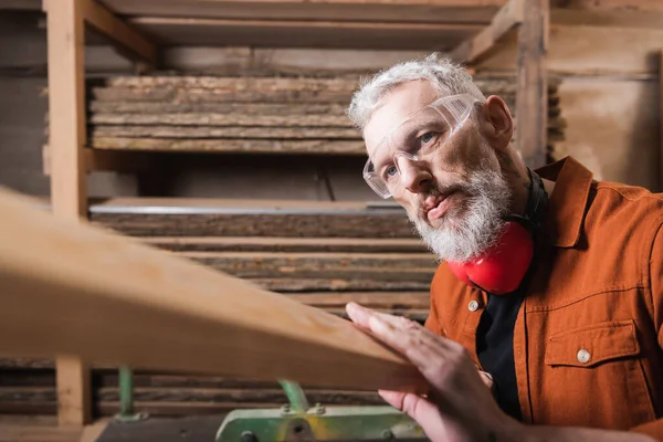 Bearded carpenter in goggles checking wooden plank in workshop — Stock Photo