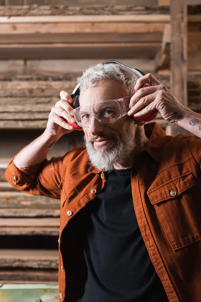 Bearded woodworker putting on protective earmuffs and looking at camera — Stock Photo