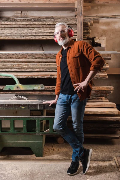 Mature carpenter standing with hand on hip near workbench with circular saw — Stock Photo