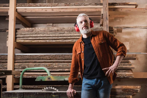 Carpenter standing with hand on hip near workbench with circular saw in workshop — Stock Photo