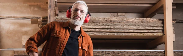 Bearded furniture designer in goggles and earmuffs looking at camera in workshop, banner — Stock Photo