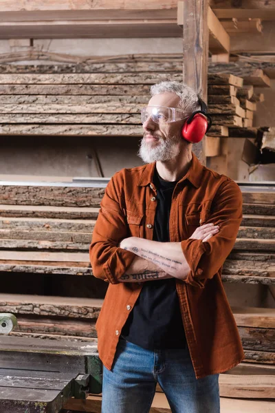 Бородатый дизайнер мебели, стоящий со скрещенными руками возле досок в мастерской — стоковое фото