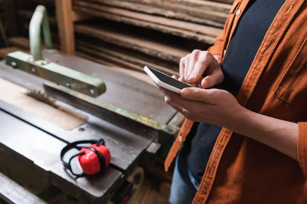 Cropped view of carpenter using smartphone with blank screen in blurred workshop — Stock Photo