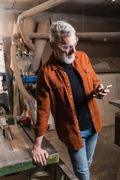 Grey haired furniture designer standing with smartphone near workbench — Stock Photo