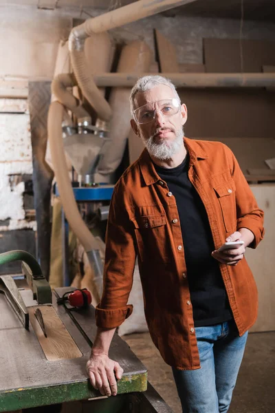 Bearded carpenter in earmuffs holding smartphone and looking at camera in workshop — Stock Photo