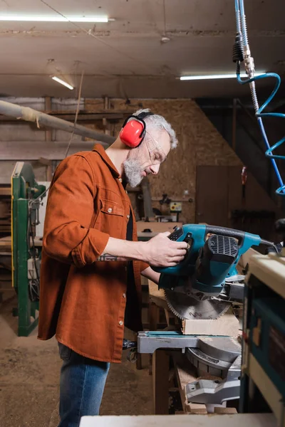 Seitenansicht des Tischlers in schützenden Ohrenschützern, der Holz auf der Gehrungssäge schneidet — Stockfoto