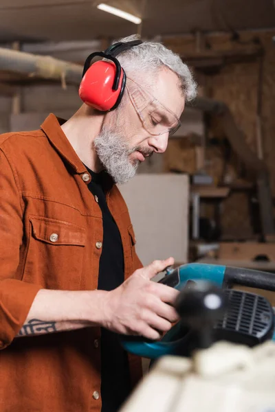 Menuisier dans des cache-oreilles protecteurs travaillant avec une scie à onglets en atelier — Photo de stock