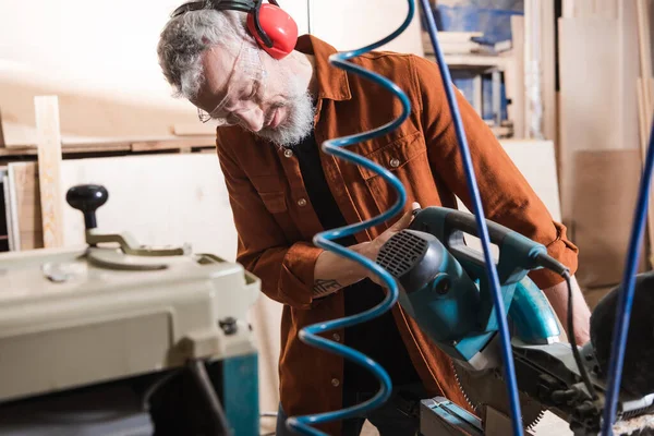 Bearded carpenter in protective earmuffs working with miter saw in workshop — Stock Photo