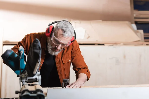 Carpenter in goggles and protective earmuffs working with miter saw — Stock Photo