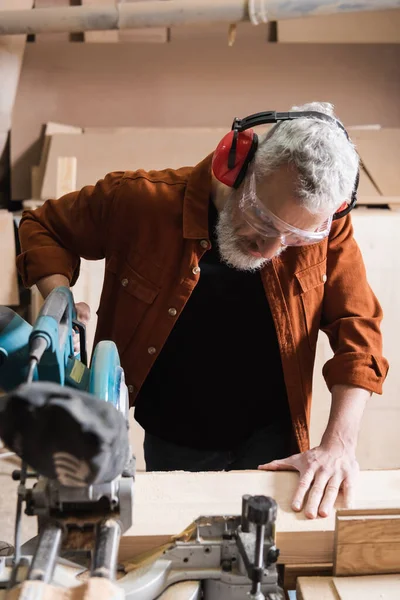 Grey haired woodworker cutting timber on miter saw in carpentry studio — Stock Photo