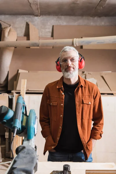 Designer de móveis maduros em óculos e auriculares de pé com as mãos em bolsos na oficina — Fotografia de Stock