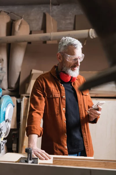 Bearded furniture designer in goggles using smartphone on blurred foreground — Stock Photo