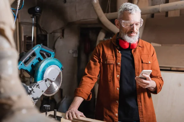 Бородатий дизайнер меблів, використовуючи мобільний телефон біля пилки — стокове фото