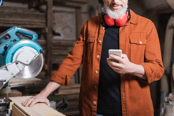 Ausgeschnittene Ansicht eines bärtigen Holzarbeiters mit Handy in der Nähe der Gehrungssäge in der Werkstatt — Stockfoto