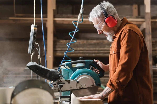 Vue latérale du charpentier barbu coupant le bois sur la scie à onglets — Photo de stock