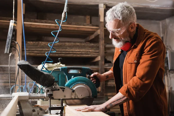 Travailleur du bois mature dans des lunettes de coupe de bois avec scie à onglets — Photo de stock