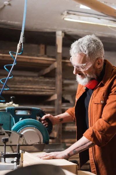 Tätowierter Schreiner mit Brille sägt Holz in Holzwerkstatt — Stockfoto