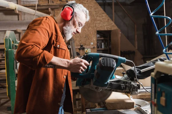 Grauhaariger Schreiner arbeitet in Holzwerkstatt mit Gehrungssäge — Stockfoto